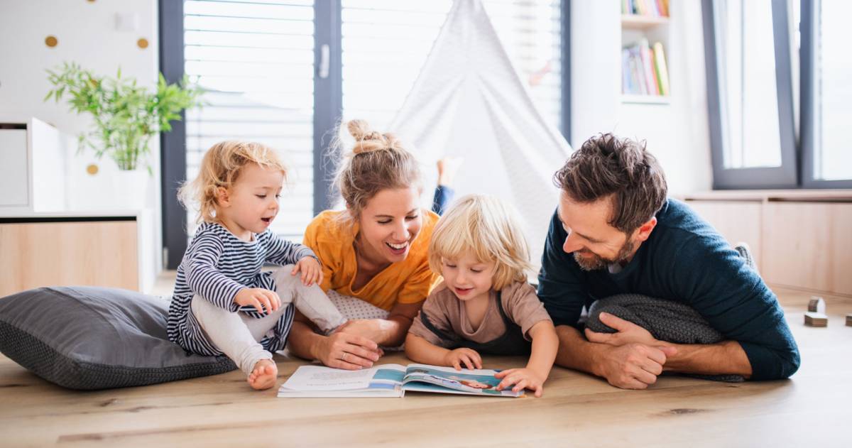 Jong gezin met twee kleine kinderen binnenshuis in de slaapkamer die een boek lezen.